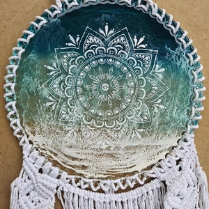 Op de afbeelding: Een witte macramé droomvanger met een ronde, turquoise en witte hars mandala. De mandala heeft een wit ontwerp op een turquoise achtergrond met een witte rand.