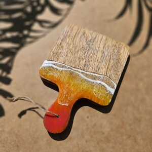 May include: A small wooden cutting board with a colorful resin inlay that resembles a sunset over the ocean. The board has a hole for hanging and a red, orange, and yellow resin design.