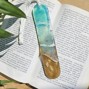Op de afbeelding: Een handgemaakte hars boekenlegger met een strandscene. De boekenlegger is transparant en heeft een blauwe zee met witte golven die breken op een zandstrand. Een schelp is zichtbaar in het zand.