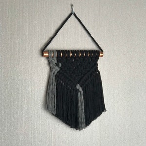 Peut inclure: Une suspension murale en macramé noire et grise avec un tuyau en cuivre en haut. La suspension a la forme d'un triangle avec des franges en bas.