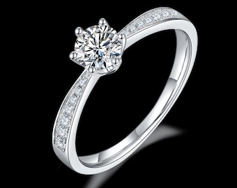 May include: A silver engagement ring with a large, round, clear gemstone at the center. The band is also silver and features smaller, clear gemstones set along the top half of the ring. The ring is set against a black background.