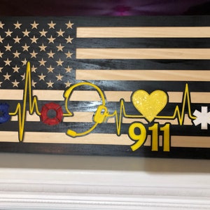 May include: Wooden American flag with a black and yellow heart, a police badge, a fire department badge, a yellow headset, and a medical star with a heartbeat line and the number 911.