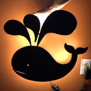 May include: A black whale-shaped night light with a white cord and a switch. The light is on and illuminating the wall behind it.