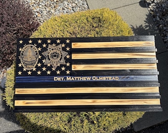 Puede incluir: Una placa de pared rectangular de madera diseñada como una bandera estadounidense estilizada con rayas alternas negras y de madera natural. El cantón presenta una insignia de la policía de Los Ángeles y una insignia del sheriff del condado de King. Una fina línea azul lleva el texto DET. MATTHEW OLMSTEAD.