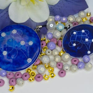 May include: Two circular blue resin charms featuring white line art and embedded clear crystals, surrounded by a collection of loose beads in shades of lavender, white, and metallic gold, with a purple flower petal visible in the background.
