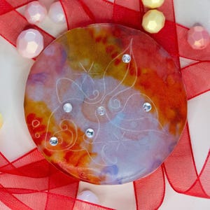May include: A circular resin disc featuring a vibrant watercolor-style blend of orange, yellow, and purple hues. The surface is etched with a delicate white line drawing of a butterfly and accented with five small, clear, sparkling rhinestones.