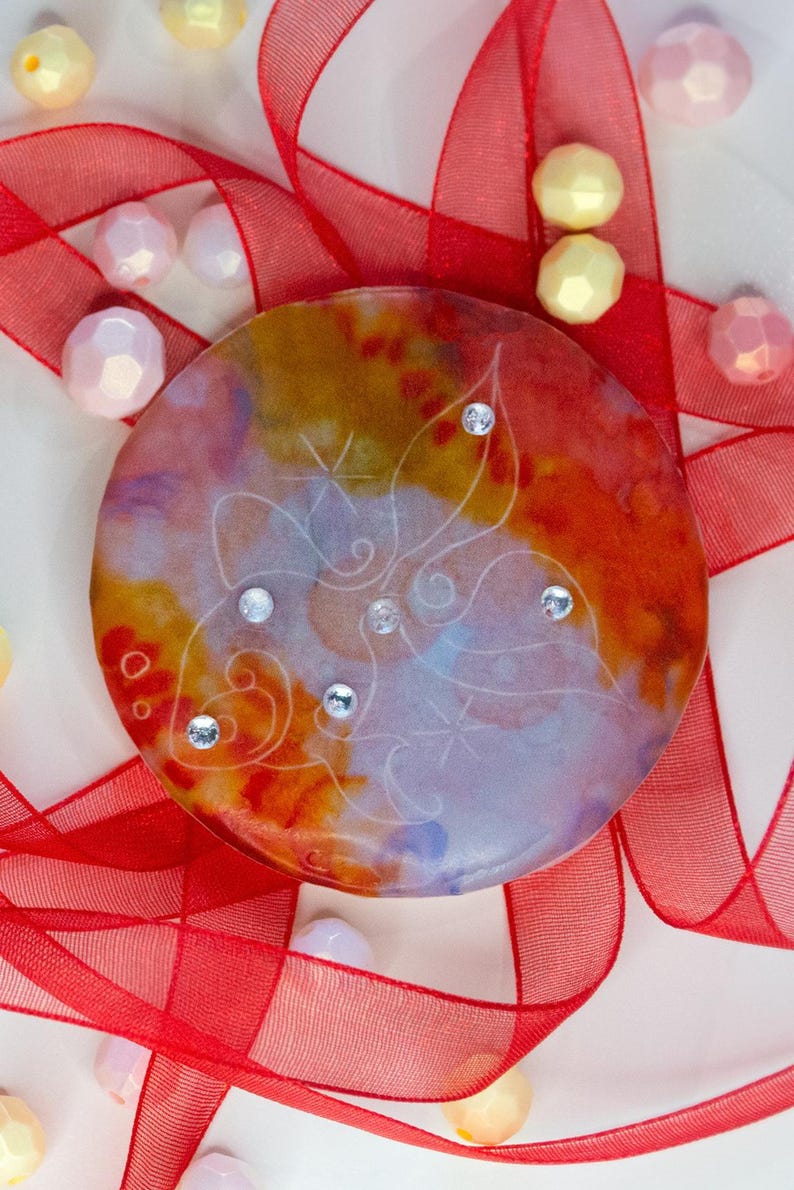 May include: A circular resin disc featuring a vibrant watercolor-style blend of orange, yellow, and purple hues. The surface is etched with a delicate white line drawing of a butterfly and accented with five small, clear, sparkling rhinestones.