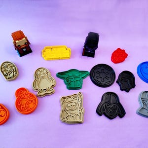 May include: A collection of 14 cookie cutters featuring characters and symbols from the Star Wars universe. The cutters are made of plastic and come in a variety of colors, including black, blue, green, red, and yellow. The cutters are arranged on a light purple surface.