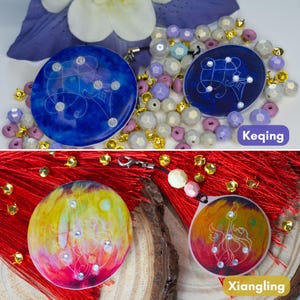 May include: Two circular charms featuring blue resin with white line art and embedded crystals, labeled Keqing, and two circular charms featuring yellow and red resin with white line art and embedded crystals, labeled Xiangling, with beads.