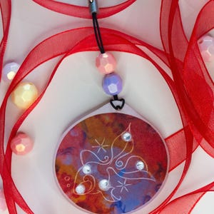 May include: A circular pendant featuring a warm-toned abstract background with a white line-art butterfly design and four small clear rhinestones. It hangs from a black cord with one peach and one lavender bead, attached to a silver-tone lobster clasp.