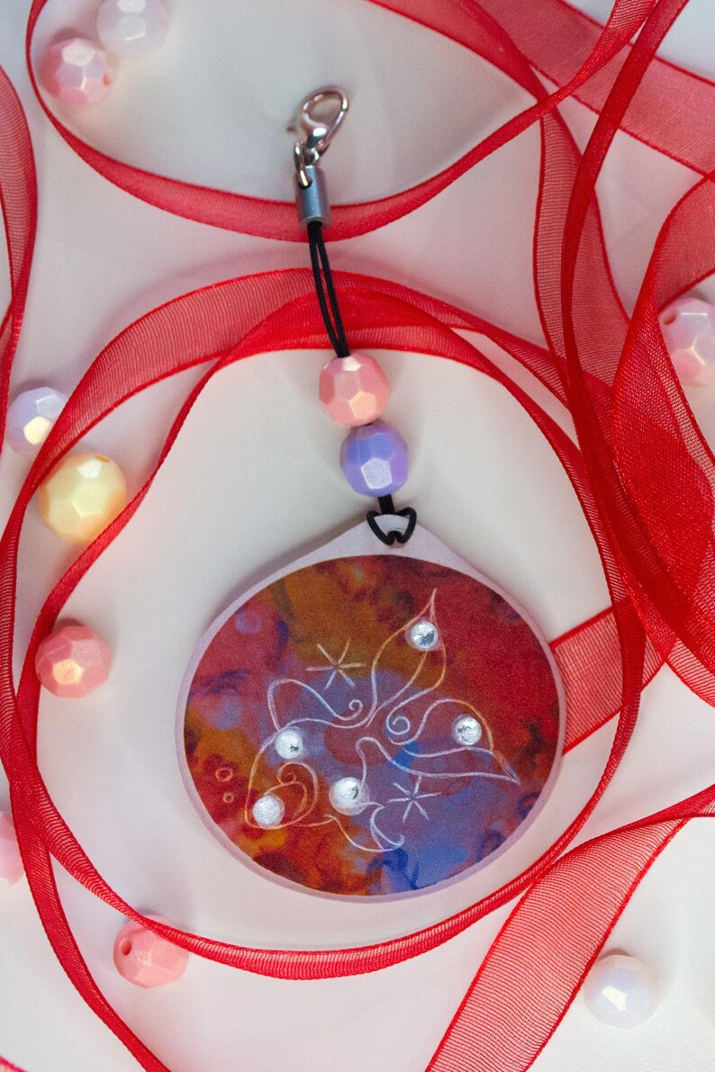 May include: A circular pendant featuring a warm-toned abstract background with a white line-art butterfly design and four small clear rhinestones. It hangs from a black cord with one peach and one lavender bead, attached to a silver-tone lobster clasp.