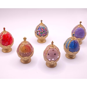 May include: Six egg-shaped ornaments with gold-colored bases and pointed finials. Each ornament features a unique color scheme including red, orange-yellow, multi-colored confetti, light pink, blue, and purple, all with a glittery finish.