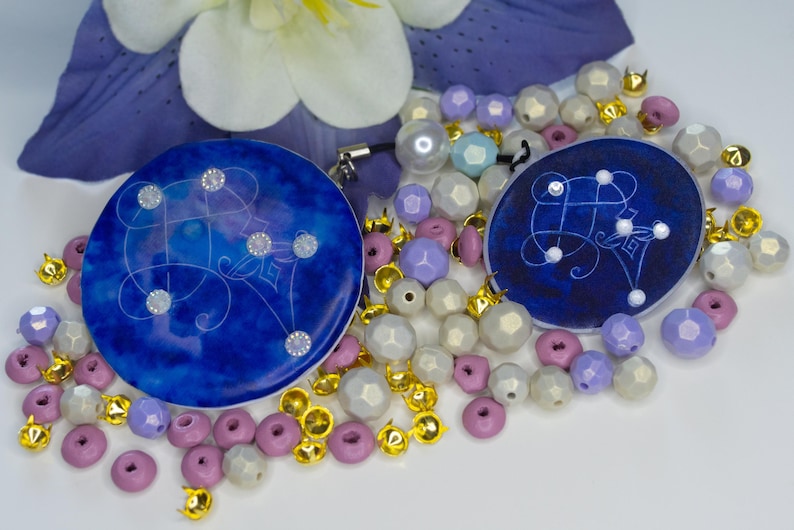 May include: Two circular blue resin charms featuring white line art and embedded clear crystals, surrounded by a collection of loose beads in shades of lavender, white, and metallic gold, with a purple flower petal visible in the background.