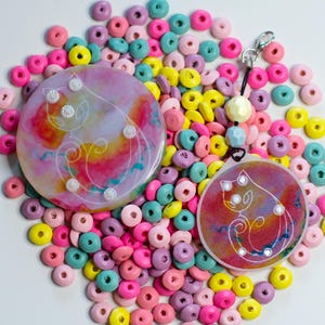 May include: Two circular resin ornaments featuring a white line art cat illustration over a vibrant pink, yellow, and blue watercolor background. The smaller ornament includes a black cord, a silver lobster clasp, and two pastel beads.
