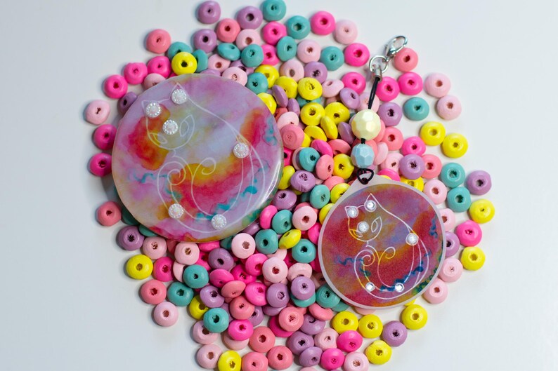 May include: Two circular resin ornaments featuring a white line art cat illustration over a vibrant pink, yellow, and blue watercolor background. The smaller ornament includes a black cord, a silver lobster clasp, and two pastel beads.