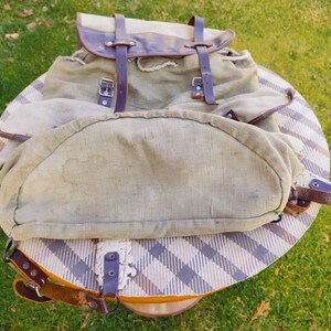 Op de afbeelding: Een vintage canvas rugzak met bruine leren banden en gespen. De rugzak heeft een groot hoofdvak en een kleiner voorvak. De rugzak is in goede staat, met enige slijtage.