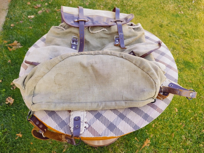 Op de afbeelding: Een vintage canvas rugzak met bruine leren banden en gespen. De rugzak heeft een groot hoofdvak en een kleiner voorvak. De rugzak is in goede staat, met enige slijtage.