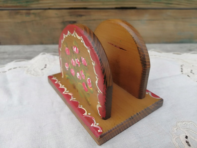 May include: A wooden napkin holder with a hand-painted design of pink roses and green leaves. The holder is painted in shades of brown, red, and white.