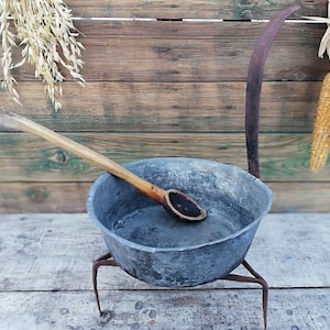 Peut inclure: Une casserole en métal gris avec une cuillère en bois à l'intérieur, posée sur un trépied en métal sur une surface en bois. La casserole est rustique et a un aspect usé.