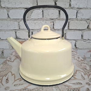Puede incluir: Hervidor de esmalte color crema con asa y borde negros. El hervidor tiene un cuerpo redondeado, un pico y una tapa con un pequeño asa de bucle. El fondo es una pared de ladrillos blancos. Un artículo de cocina vintage.