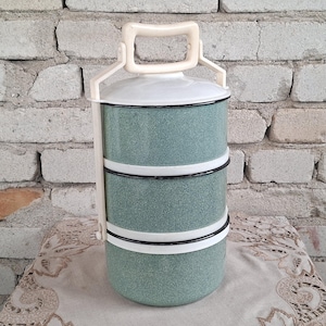 May include: A vintage three-tiered lunch container. The container is a speckled teal color with white and black trim. It has a white handle and lid. The lunchbox is sitting on a decorative cloth in front of a brick wall.