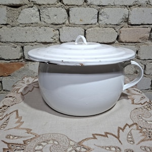 Può includere: Vaso da notte in smalto bianco con coperchio e manico. Il vaso è rotondo e poggia su un panno decorativo. Lo sfondo è un muro di mattoni. Articolo vintage.