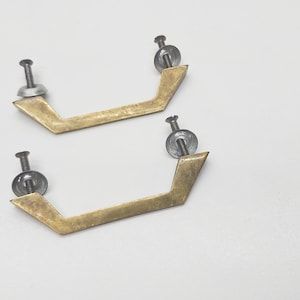 May include: Two brass cabinet pulls with a geometric shape and silver screws. The pulls are on a white background.
