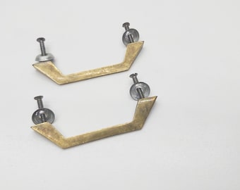 May include: Two brass cabinet pulls with a geometric shape and silver screws. The pulls are in a white background.