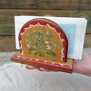 May include: A wooden napkin holder with a hand-painted design of blue flowers on a yellow background. The holder has a red trim and is decorated with a scalloped edge.