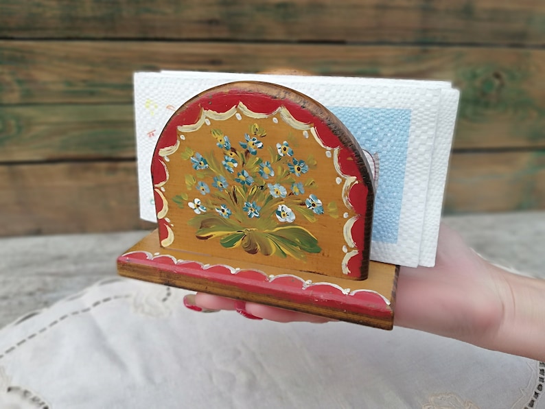 May include: A wooden napkin holder with a hand-painted design of blue flowers on a yellow background. The holder has a red trim and is decorated with a scalloped edge.