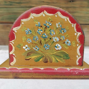 May include: A wooden napkin holder painted with a bouquet of blue and white flowers on a yellow background. The holder has a red trim with white scalloped edges.
