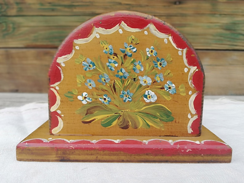 May include: A wooden napkin holder painted with a bouquet of blue and white flowers on a yellow background. The holder has a red trim with white scalloped edges.