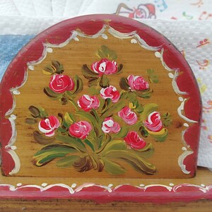 May include: A wooden napkin holder with a hand-painted design of pink roses and green leaves on a brown background. The holder has a red base and a red top with white scalloped edges.