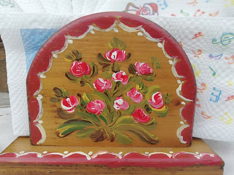 May include: A wooden napkin holder with a hand-painted design of pink roses and green leaves on a brown background. The holder has a red base and a red top with white scalloped edges.