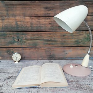 May include: A white and pink vintage desk lamp with a flexible arm, a book, and a small clock on a wooden table.