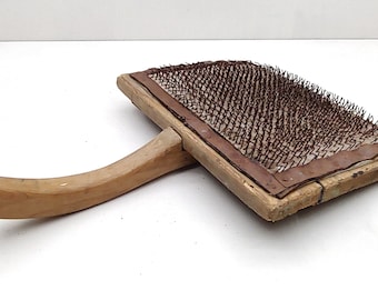 Könnte beinhalten: Ein hölzerner Wollkarden mit einem langen Holzgriff. Die Kardenfläche ist mit Metallzähnen bedeckt.