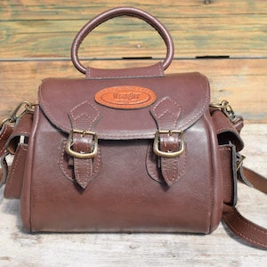 May include: A brown leather satchel with a top handle and a shoulder strap. The bag features two front buckles and a small leather patch with the "Wrangler" logo. The bag has two side pockets and a circular handle.