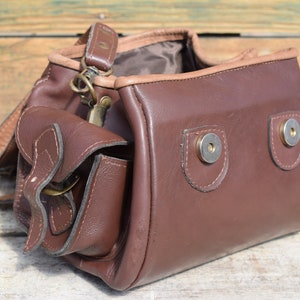 May include: A brown leather shoulder bag with a flap pocket and a long strap. The bag has a rectangular shape with rounded edges and metal snap closures. The bag is made of a smooth, dark brown leather.