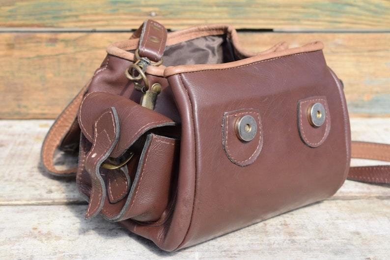 May include: A brown leather shoulder bag with a flap pocket and a long strap. The bag has a rectangular shape with rounded edges and metal snap closures. The bag is made of a smooth, dark brown leather.