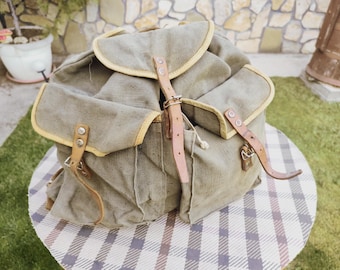 Könnte beinhalten: Ein Vintage-Rucksack aus Canvas mit Lederriemen und Schnallen. Der Rucksack ist hellgrün mit gelbem Besatz.