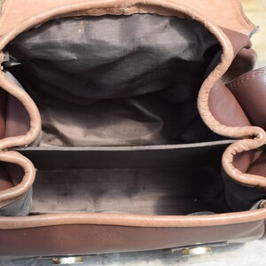 May include: Open view of a brown leather bag with a flap closure. The bag has a rectangular shape with a lighter brown trim and interior. Metal hardware includes rings and clasps. The bag appears to be empty.