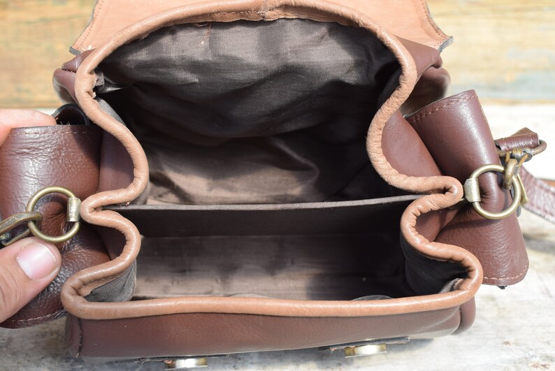 May include: Open view of a brown leather bag with a flap closure. The bag has a rectangular shape with a lighter brown trim and interior. Metal hardware includes rings and clasps. The bag appears to be empty.