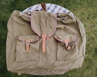Könnte beinhalten: Ein großer, brauner Canvas-Rucksack mit Lederriemen und Schnallen. Der Rucksack hat zwei große Taschen auf der Vorderseite und eine kleinere Tasche oben.