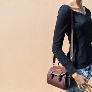 May include: A brown leather crossbody bag with a top handle and a shoulder strap. The bag has two front buckles and a small, round emblem on the front. The bag is worn by a mannequin wearing a black long-sleeve shirt and blue jeans.