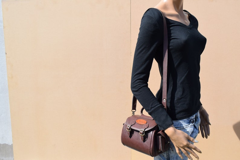May include: A brown leather crossbody bag with a top handle and a shoulder strap. The bag has two front buckles and a small, round emblem on the front. The bag is worn by a mannequin wearing a black long-sleeve shirt and blue jeans.