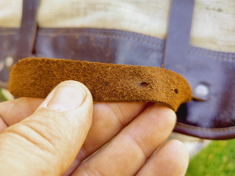 Op de afbeelding: Close-up van een bruine leren riem met twee gaten. De riem is bevestigd aan een groter stuk leer.