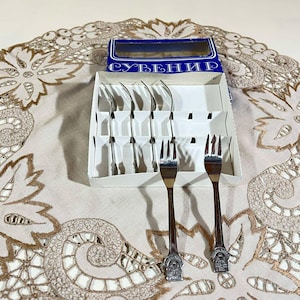 Può includere: Un set di sei forchette souvenir color argento in una scatola bianca con scritta blu che dice "СУВЕНИР". Le forchette sono disposte in file e sono pronte per essere utilizzate.