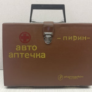 Op de afbeelding: Een bruine leren EHBO-doos met een metalen handvat en sluiting. De doos heeft een rood kruis en de tekst "авто аптечка - пирин - pharmachim BULGARIA" in gouden letters.