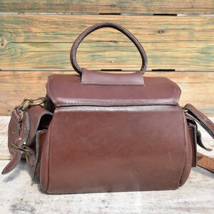 May include: A brown leather camera bag with a top handle and a shoulder strap. The bag has a rectangular shape with a flap closure and two side pockets. The hardware is brass-colored. The bag is sitting on a wooden surface.