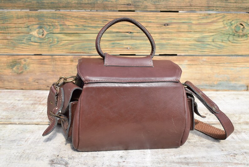 May include: A brown leather camera bag with a top handle and a shoulder strap. The bag has a rectangular shape with a flap closure and two side pockets. The hardware is brass-colored. The bag is sitting on a wooden surface.